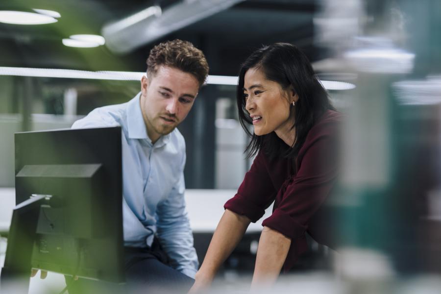 A man and a woman are leaning over computers, engaged in a discussion.