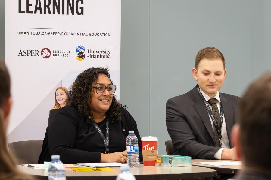 Woman sits at a table addressing a room of people, a man sits next to her