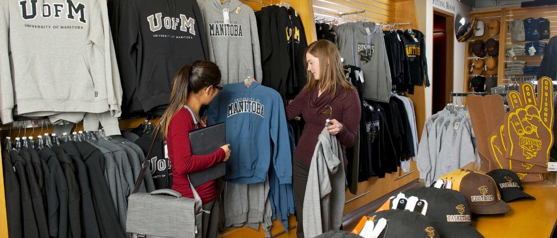 A staff member holding one grey and one blue university-branded hoodie while talking with a student in the fort garry bookstore.