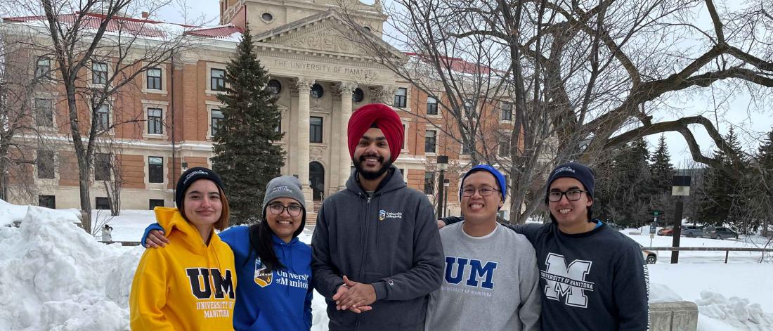 Five students pose in front of the Administration building wearing University of Manitoba branded merchandise.