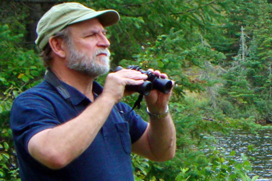 Zoologist Bill Watkins stands outdoors holding a pair of binoculars gazing out at a forested area.