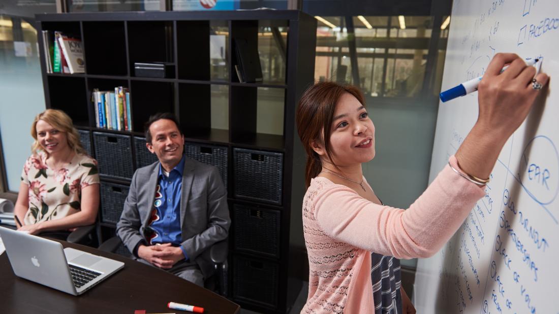 A woman writing on a whiteboard as her colleagues sit at a boardroom table.