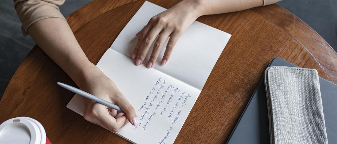 Person writing in a notebook using a blue pen, at a wooden desk.