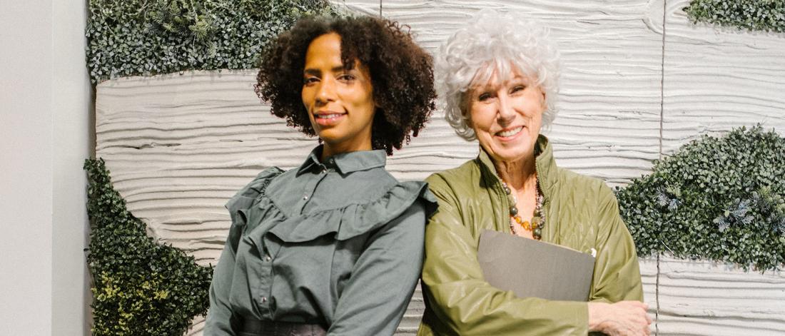 Two diverse women, who are different ages are smiling and standing back to back.