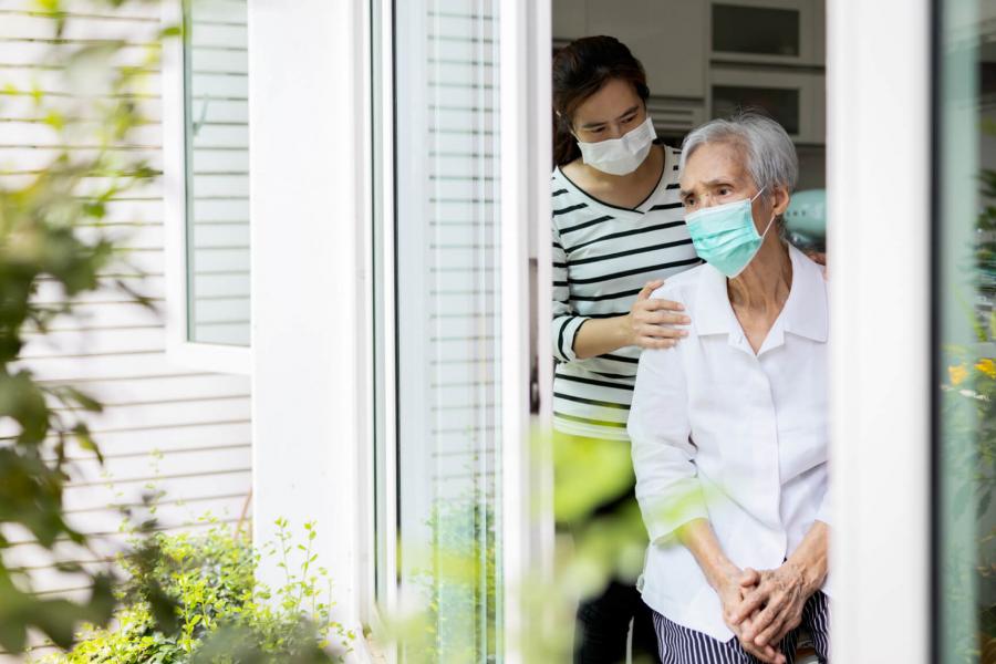 An older woman and her daughter, wearing masks look outside a window in their home.