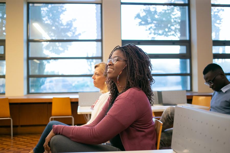 A female student is seated in a room across from her female instructor, with a male student seated behind them listening to a presentation.