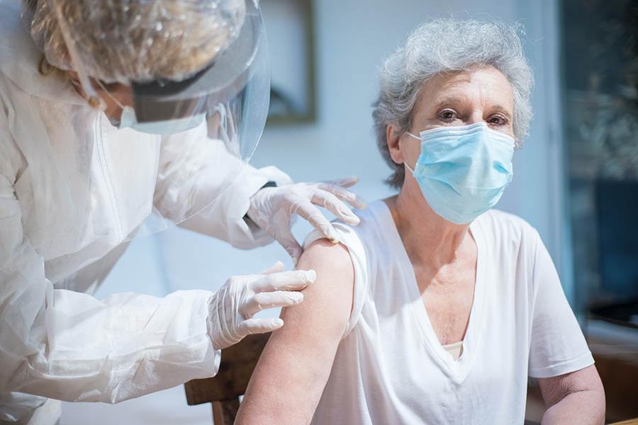 An older woman is seated and waits to receive a vaccine in her arm from a health care professional.