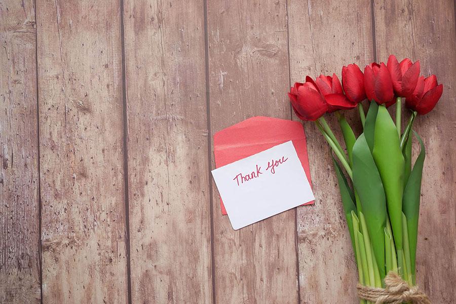 Red tulips on a wooden table. On the right side is a red envelope and a white card with thank you text written.