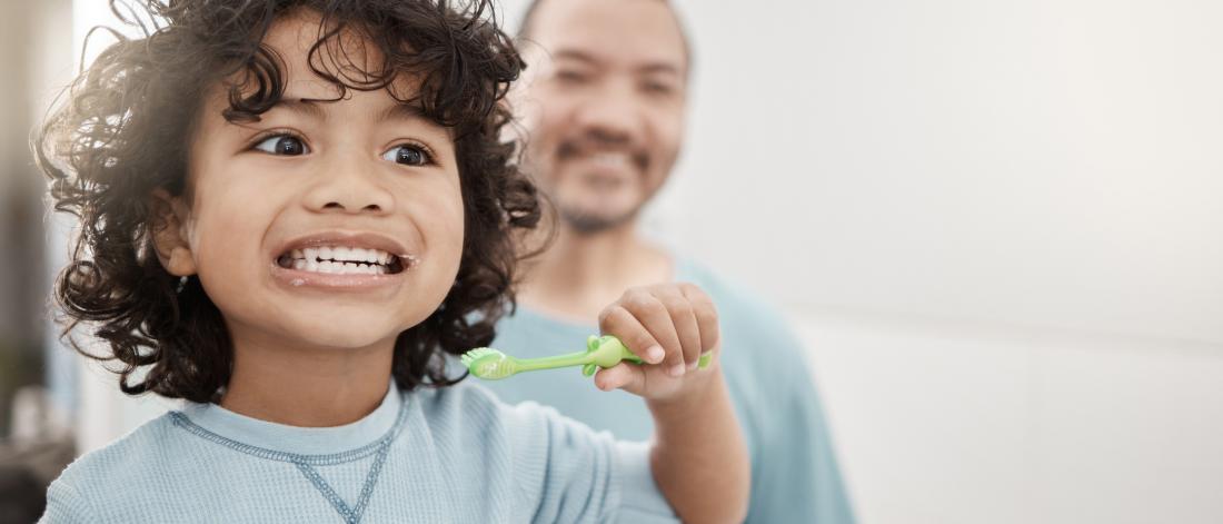 Young boy with curly hair brushes his teeth in a mirror - his father stands behind him supervising.