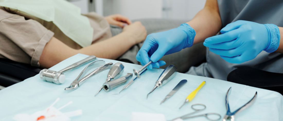 A patient reclined in an examine chair and a dentist picking up an instrument from a tray.