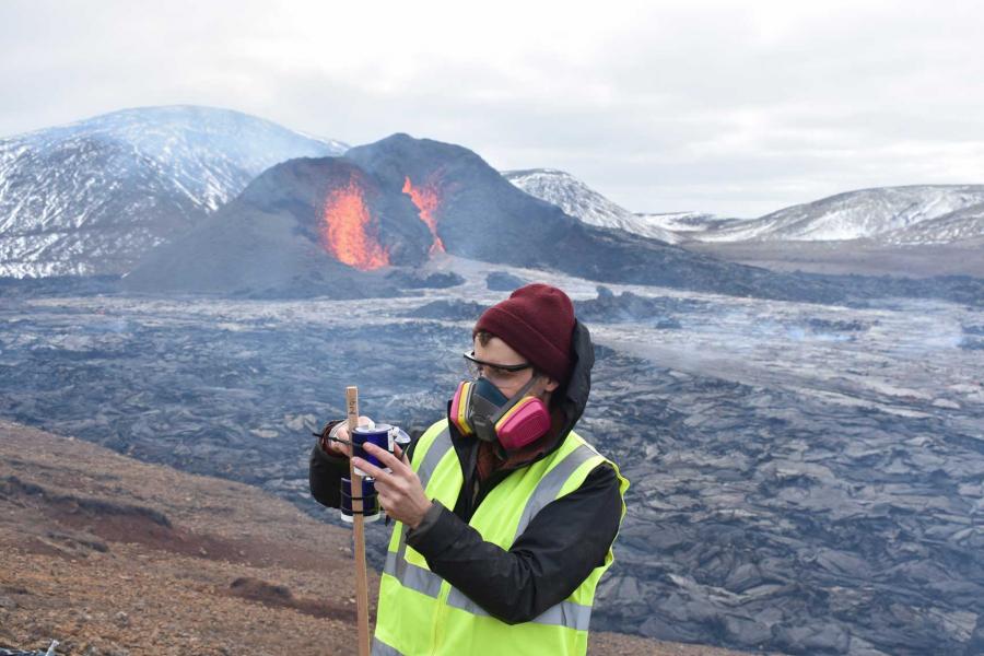 A researcher installs equipment as the main crater vent of Fagradalsfjall erupts in the background.