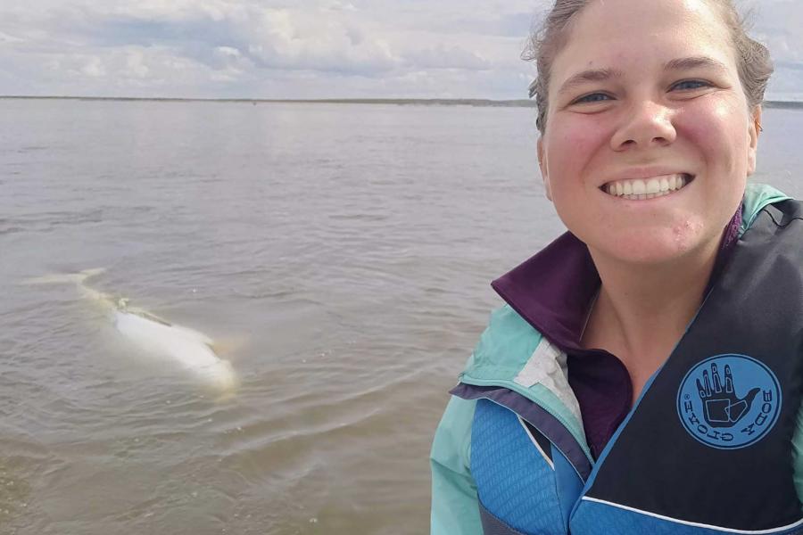 Emma Ausen smiling on a boat as a beluga surfaces the water behind her.