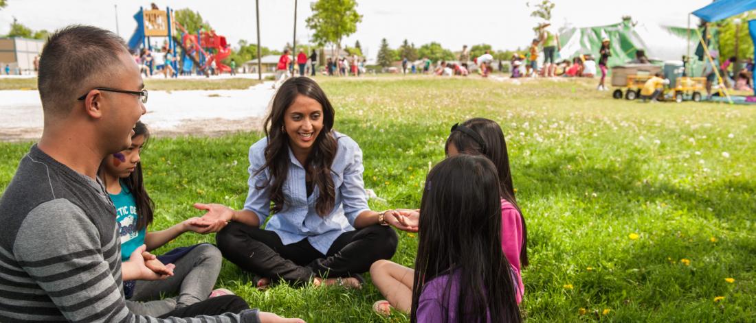 Two teachers sit on the ground in a circle with 3 young students, several other children play in the background.