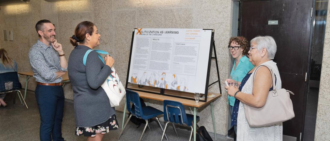 Assistant Prof. Joe Curnow, student Justin Fraser and two attendees talking at the 2019 Homecoming near a display for Politicization As Learning. 