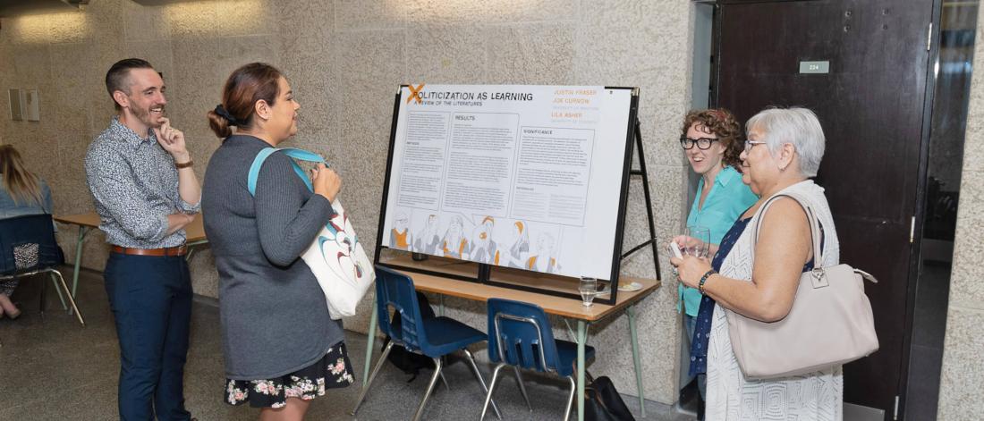 Assistant Prof. Joe Curnow, student Justin Fraser and two attendees talking at the 2019 Homecoming near a display for Politicization As Learning.