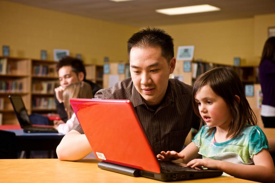 A teacher sits at a table with a young student helping her work on a laptop computer. 