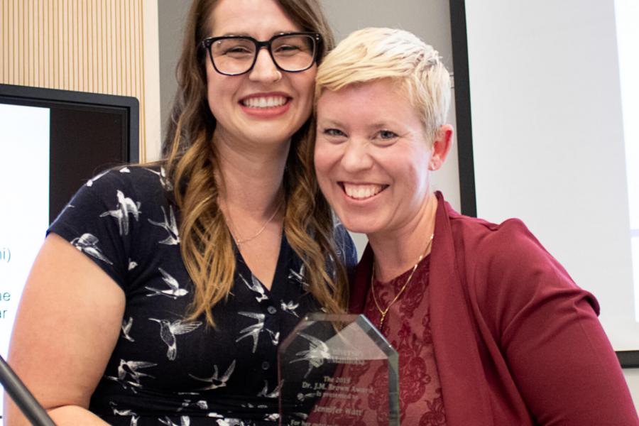 Two smiling women stand side-by-side as one receives an award. 