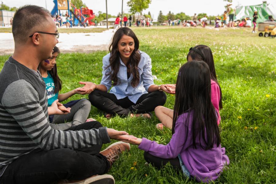 Two teachers sit on the ground in a circle with 3 young students, several other children play in the background.