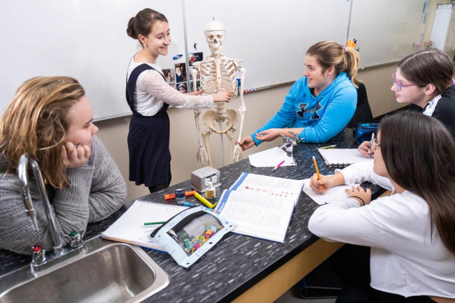 A teacher stands in front of students and points to parts of a human skeleton model.