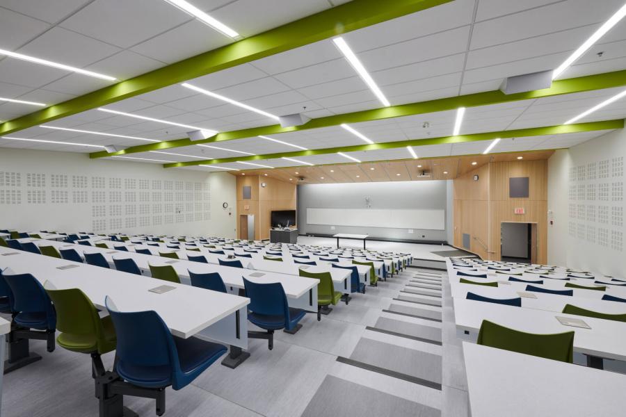 The brightly lit modern classroom space of room 290 from the back row looking towards the front of the room with rows of bright white tables and green and blue chairs.