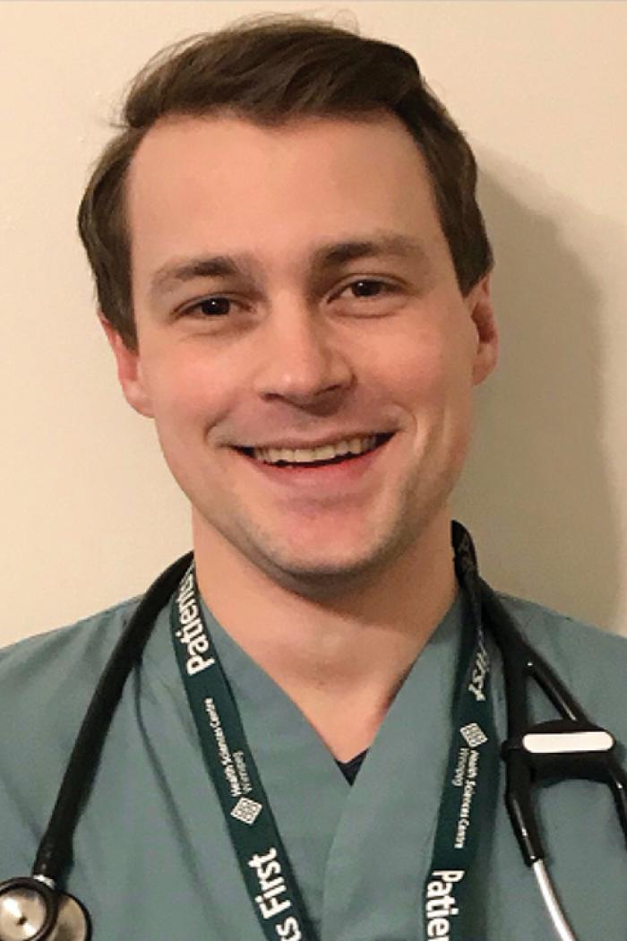 Kevin Stockwell, wearing medical scrubs with a stethoscope around his neck, stands smiling and posing for the camera. 