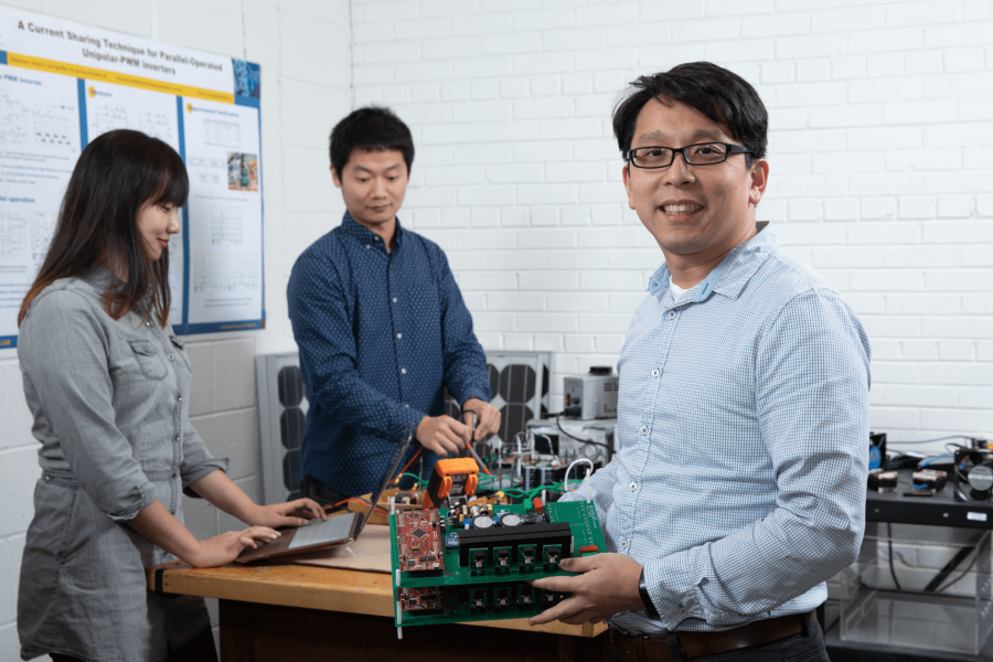 Two engineering students stand at a desk with a professor.