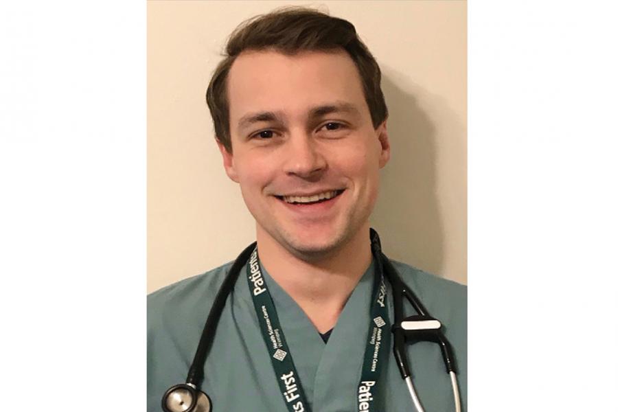 Kevin Stockwell, wearing medical scrubs with a stethoscope around his neck, stands smiling and posing for the camera. 