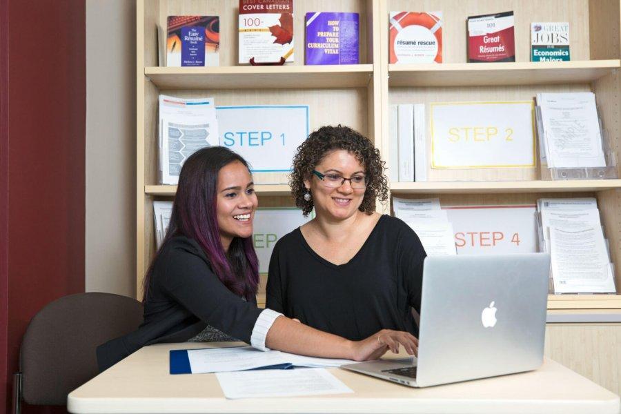 Two women sit at a table with an open laptop. Both are smiling and looking at the laptop screen.