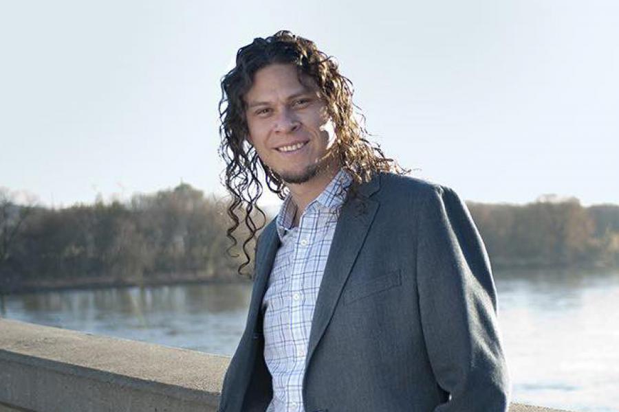 Ricardo Mantilla standing on a bridge over a river wearing a sports jacket and a button up shirt, smiling and posing for the camera. 