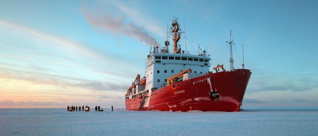 The Amundsen vessel stopped in frozen ice. Several crew members gather on the ice with three snowmobiles. 