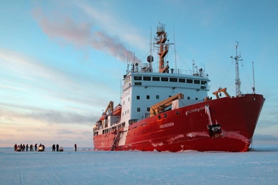 The Amundsen vessel stopped in frozen ice. Several crew members gather on the ice with three snowmobiles. 