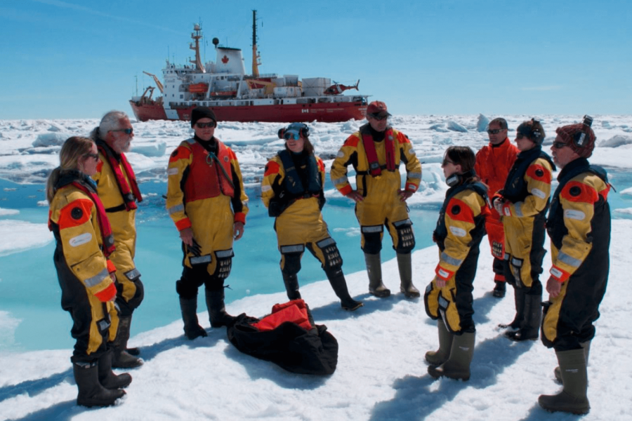 A team of researchers stand on the ice in matching yellow and orange suits. 