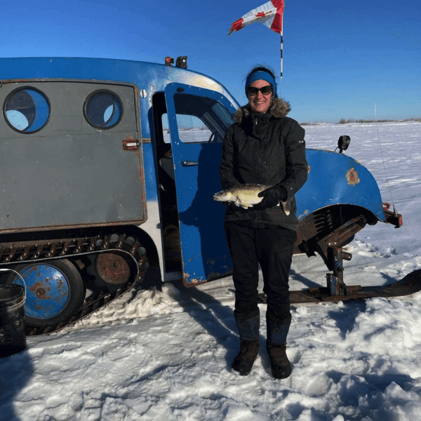 Lisa bergen standing on the ice in front of a bombadier, holding a fish.  