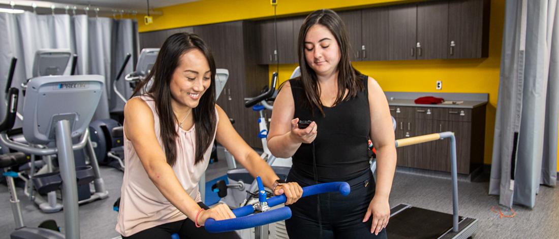 A student rides a stationary bike while another carefully monitors.