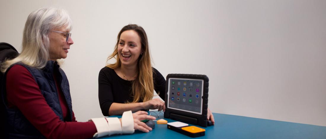 An occupational therapist sits at a blue table in a room with a patient looking at a tablet device.