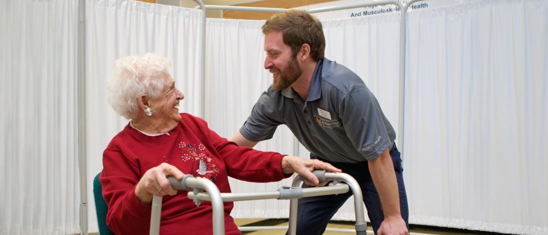 A physical therapist bends slightly to talk to an elderly patient seated with a walking aid in front of her.
