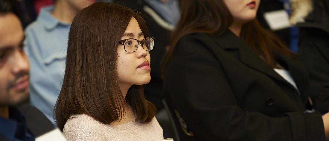 Woman with glasses sitting down listening to a presentation. She is looking at the presenter, away from the camera.