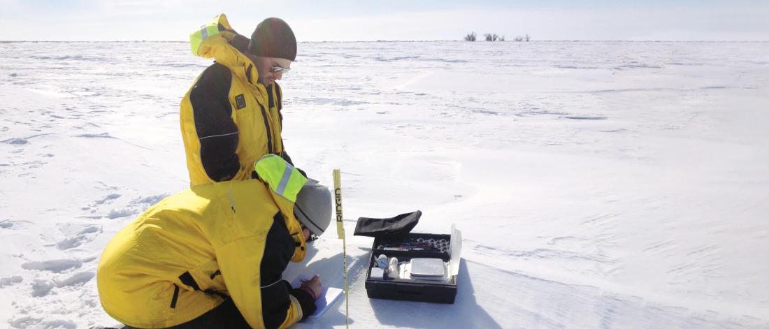 Two civil engineers outside in an open field of snow taking some measurements.