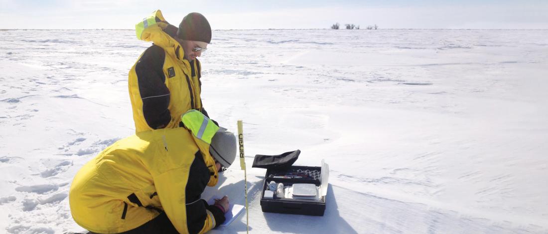 Two civil engineers outside in an open field of snow taking some measurements.