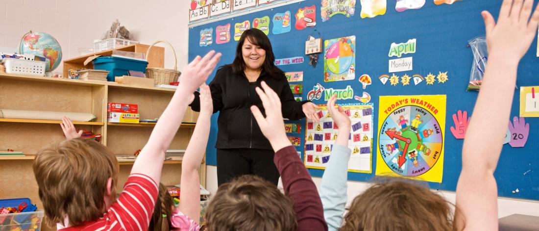 A teacher stands at the front of a classroom filled with students with raised hands.