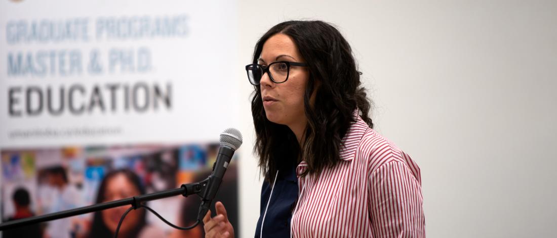 An education PhD student speaks into a microphone while standing at a podium. 