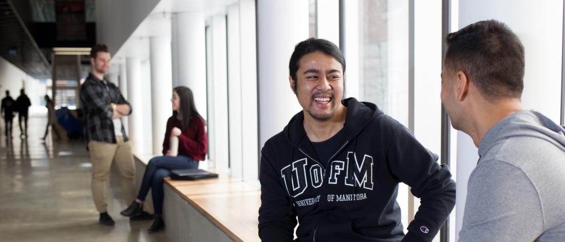 An arts student sits in a hallway and speaks with a fellow student while smiling.