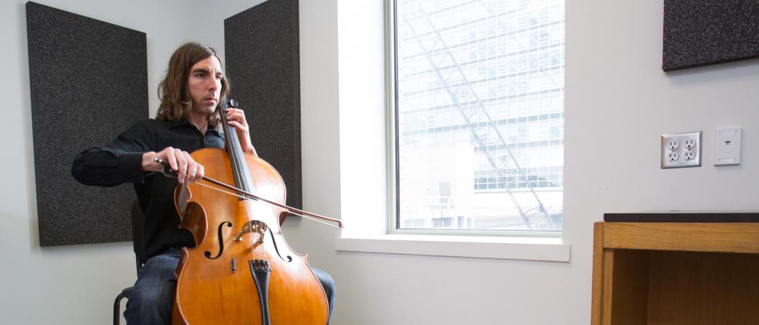 A student plays an instrument near a large window in a practice room.