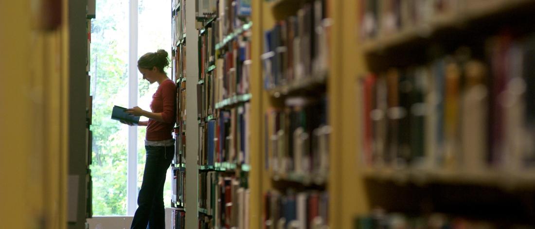 A students stands while reading a book in a library.