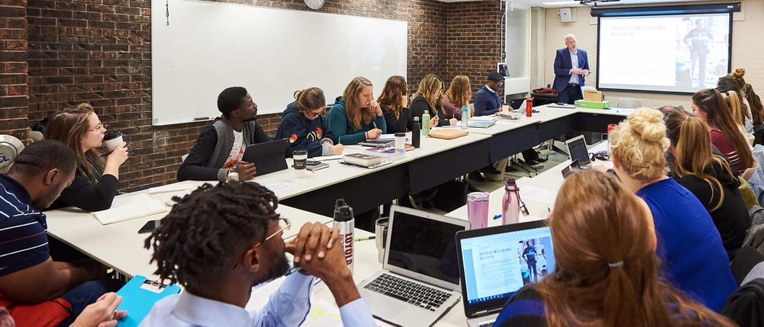 A classroom of attentive students seated in a large rectangular table watch a presentation.