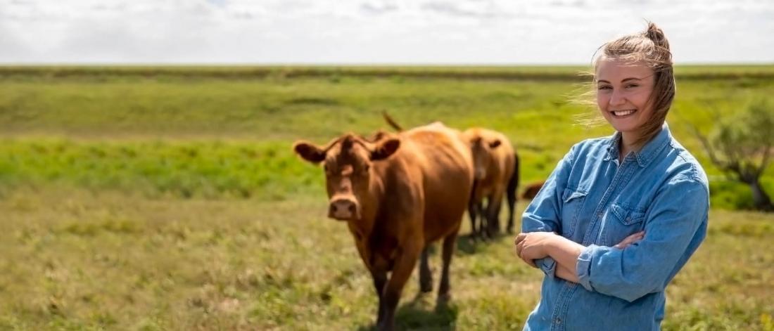 A student stands in an open field, smiling with arms crossed and several cattle walking towards her.