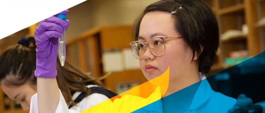 A student in a lab holds a test tube sample of a clear liquid. Another student can be seen working in the background.