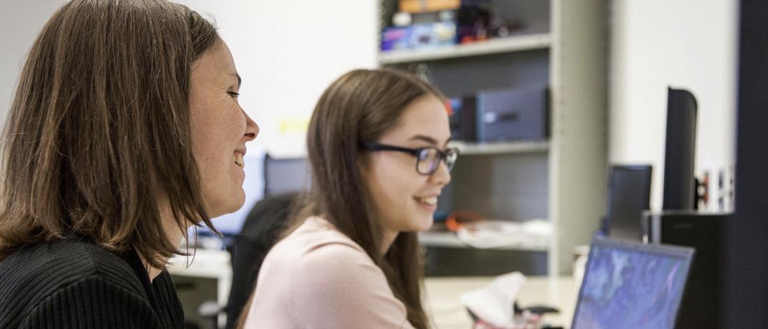 A professor sits beside a student as she works on a computer.