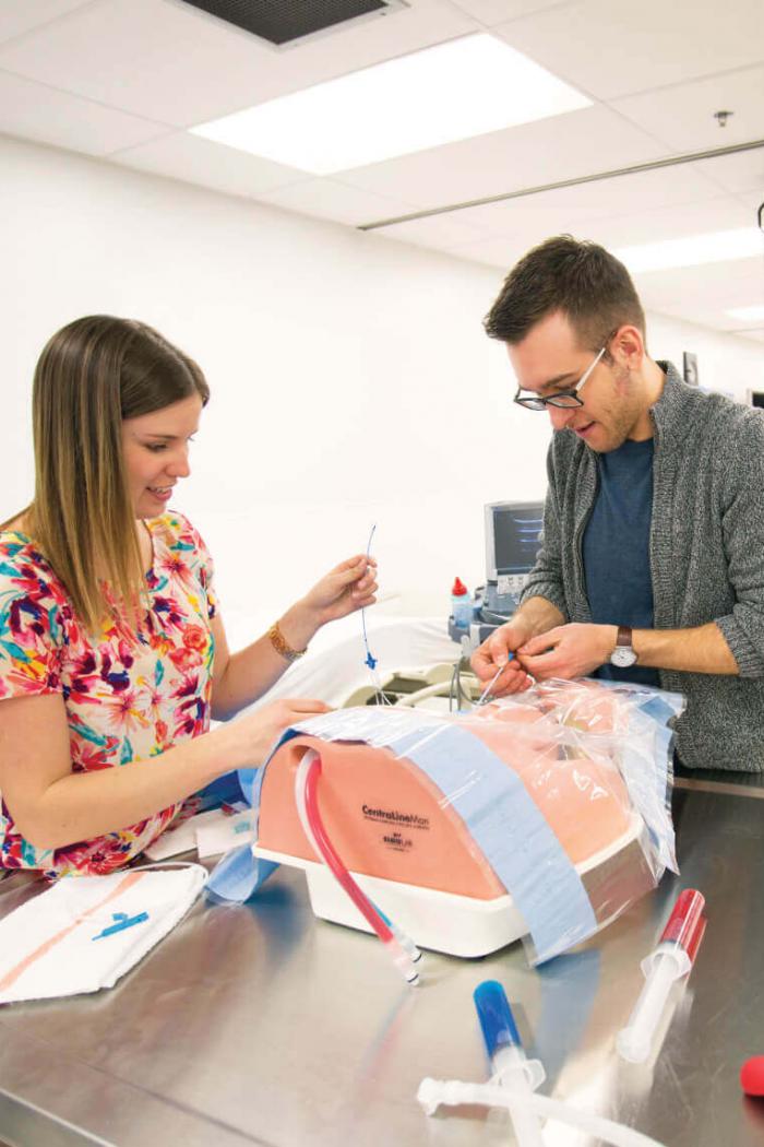 Two medical students practice administering a medical drip to a practice mannequin.