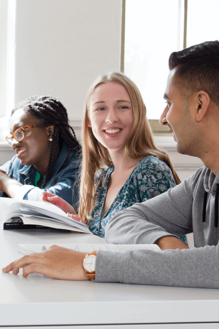 Three students seated at a desk together talking as they work.